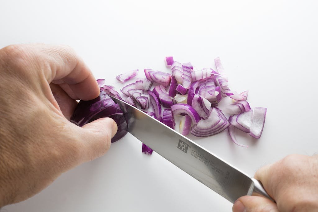 Close-up of hands slicing purple onions on a white cutting board with a sharp knife.