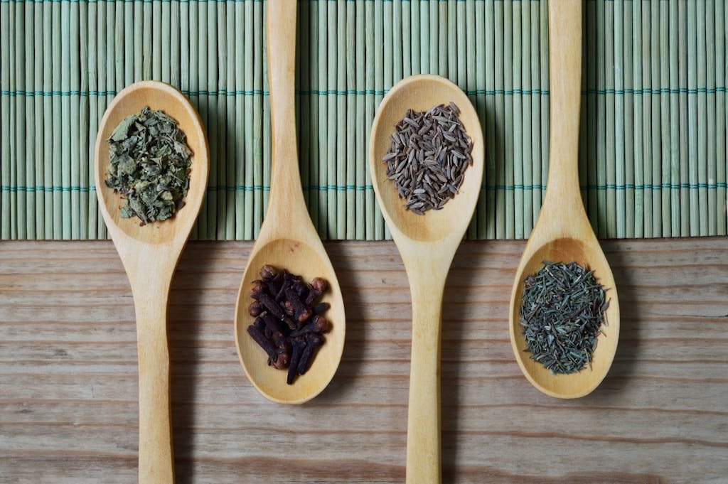 Overhead view of wooden spoons filled with spices and herbs on a bamboo mat, ideal for culinary themes.